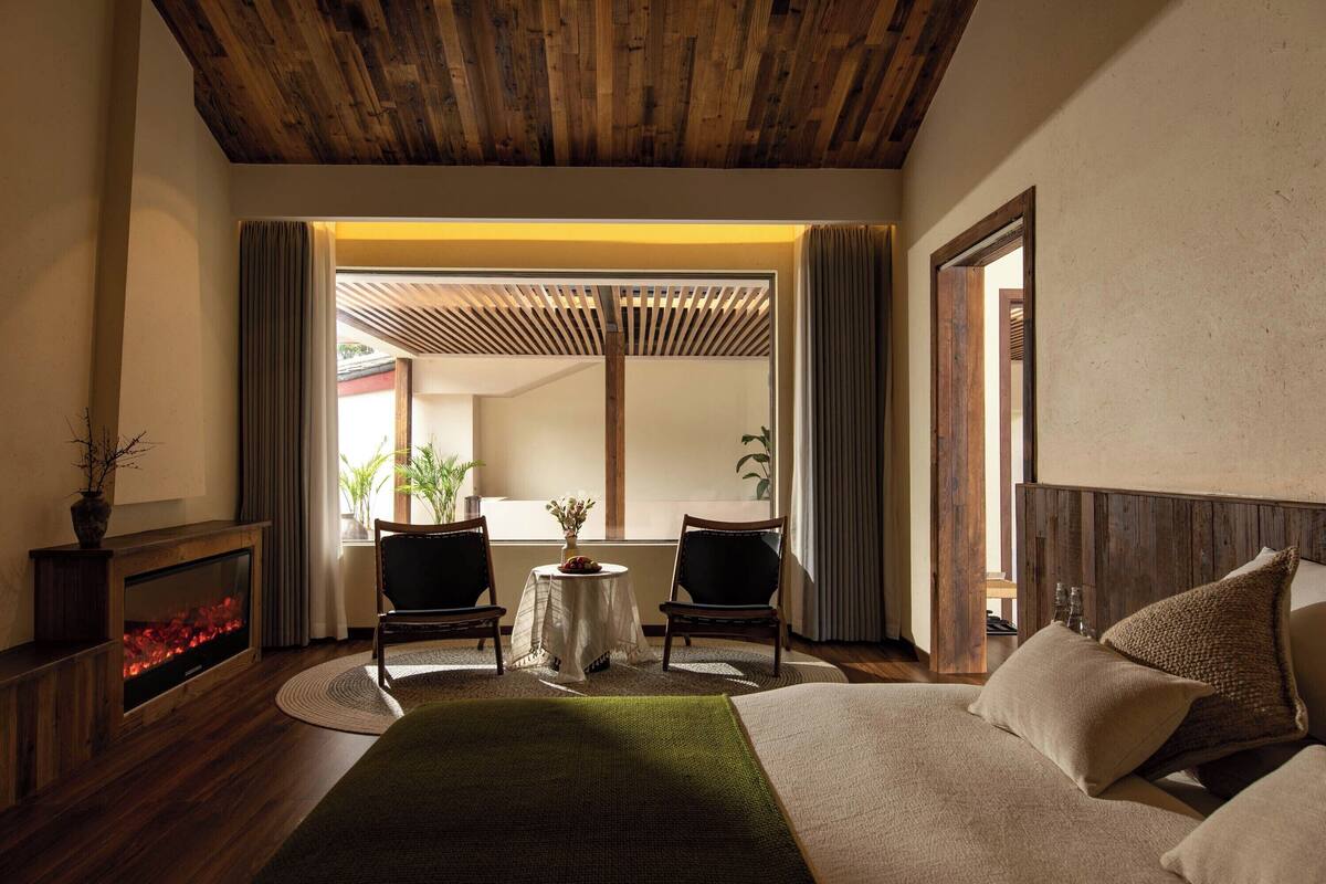 A room with a bed, two chairs, a small table, a fireplace, and a view through a large window.