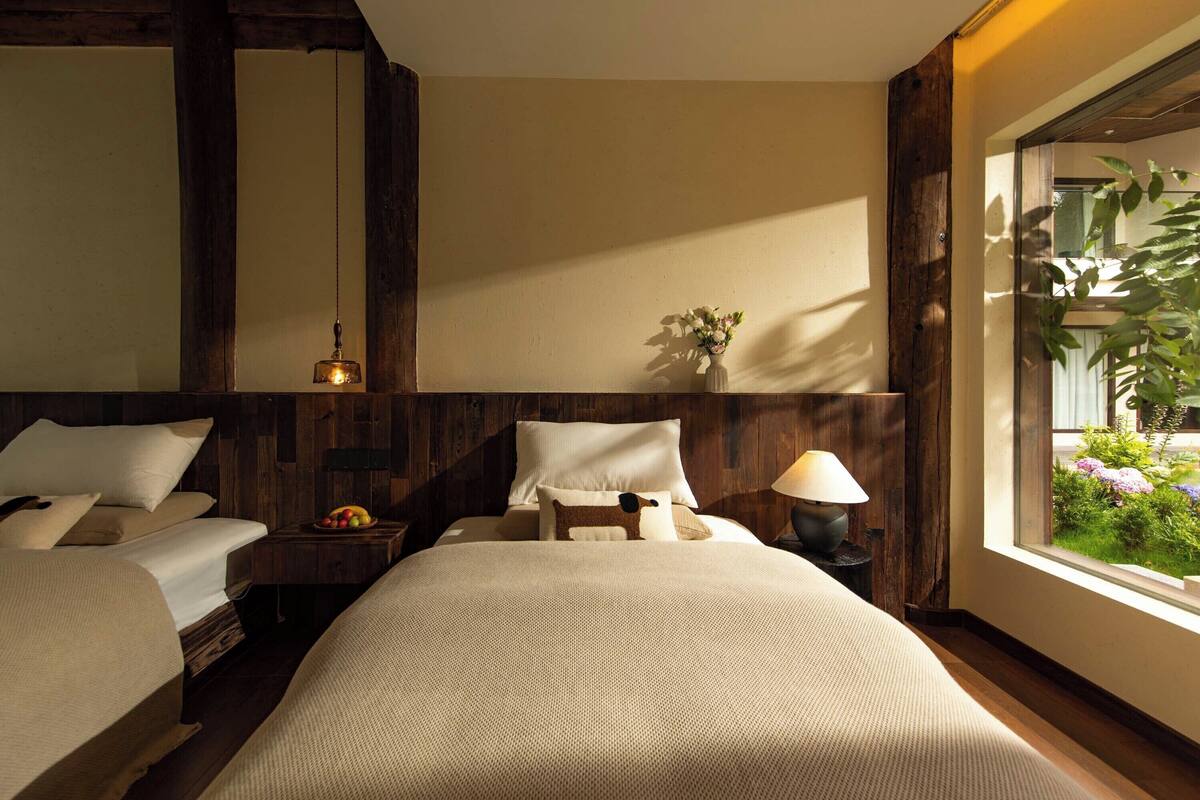 A hotel room with two beds, a nightstand with a lamp, a vase with flowers, and a window with a view of greenery.