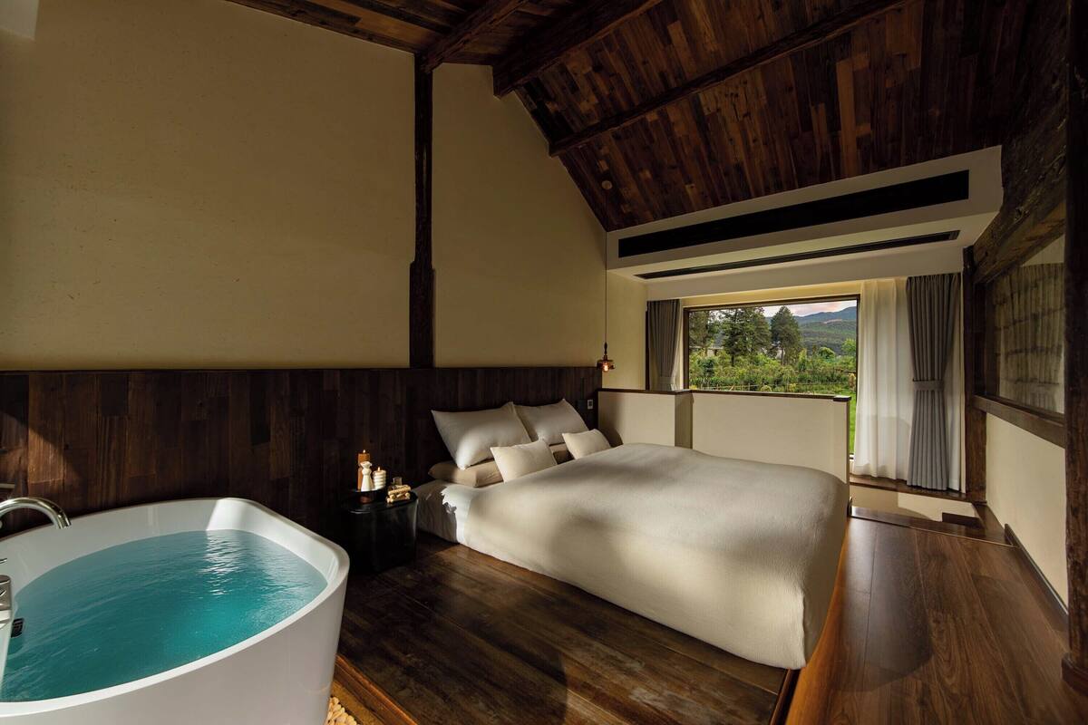 A bedroom with a large bed, a wooden ceiling, a bathtub, and a window with a view of greenery.