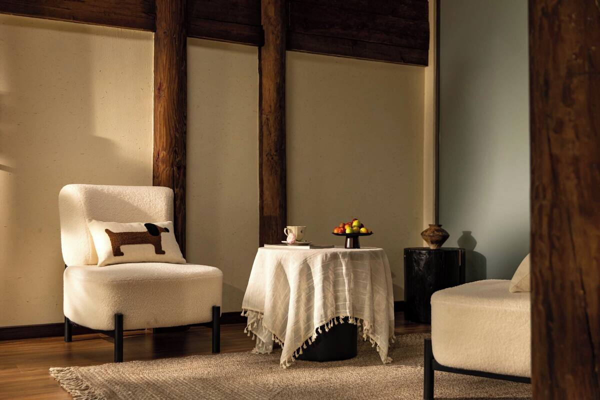 A cozy room with a wooden ceiling, a white armchair with a dog pillow, a small table with a fruit bowl, and a vase.