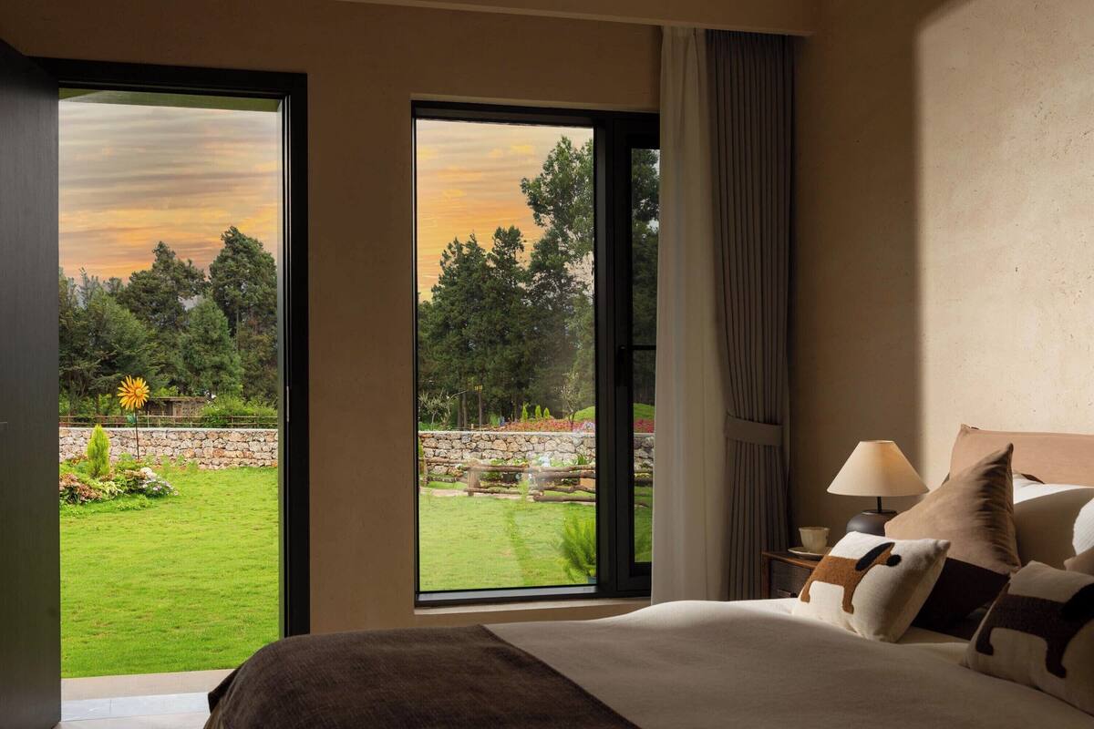 A bedroom with a large window, a bed with multiple pillows, a nightstand with a lamp, and a view of a garden and trees.