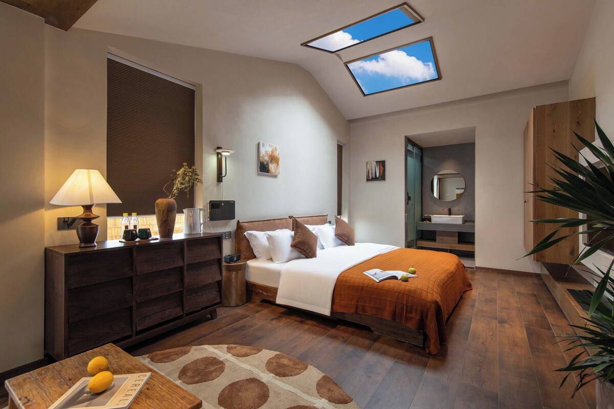Une chambre moderne avec une lucarne, un lit recouvert d’une couverture marron, une commode en bois et une petite table sur laquelle sont posés des citrons.