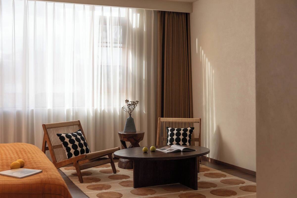 Une chambre d’hôtel avec un lit, deux chaises, une petite table ronde et une fenêtre avec des rideaux légers.