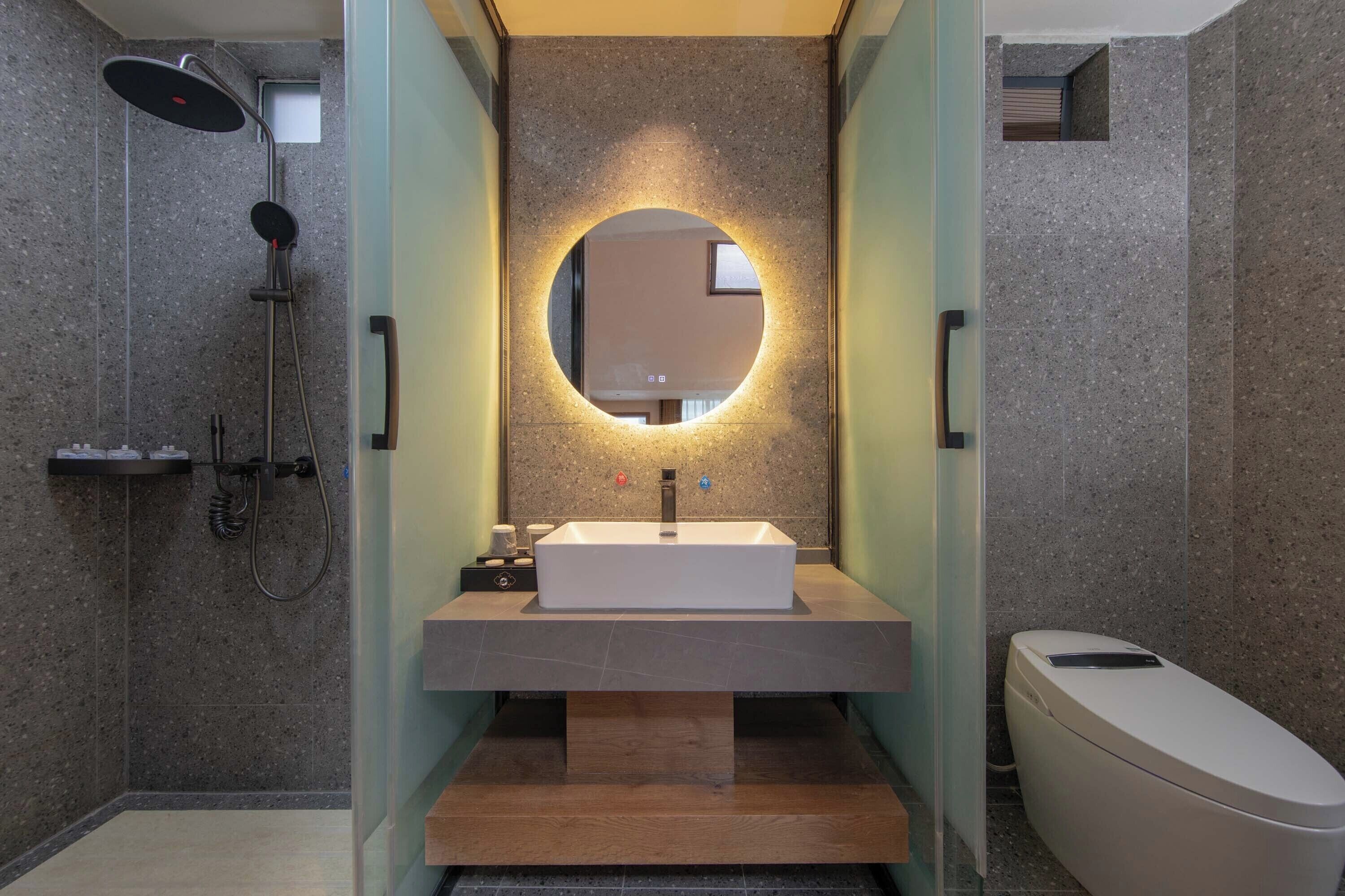 Ein modernes Badezimmer mit freistehender Waschtisch, einem runden Spiegel mit integrierter Beleuchtung, einer Handbrause und einer Toilette.