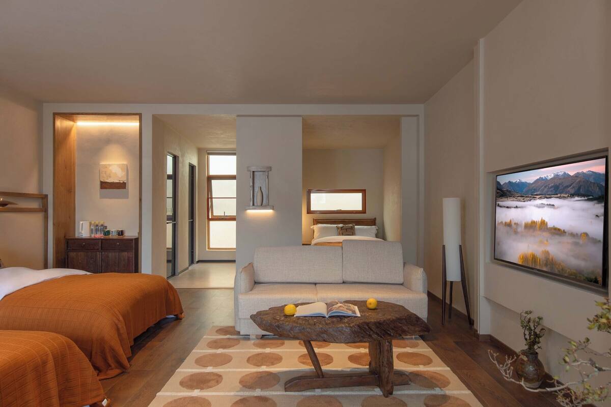 Une chambre d’hôtel avec deux lits, un canapé, une table basse avec des livres et des citrons, un téléviseur et un tableau représentant un paysage.