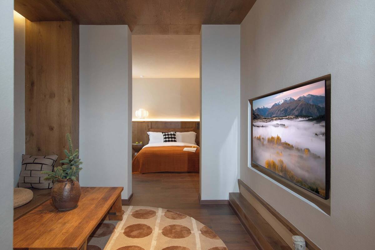 Une chambre d’hôtel avec un lit, une table de chevet ornée d’un vase, une grande fenêtre donnant sur les montagnes et un banc en bois.