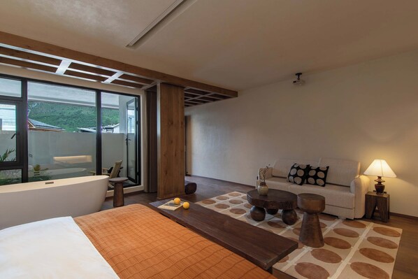 Habitación de hotel con una cama grande, un sofá, una mesa de centro y vistas a la montaña a través de amplias ventanas.