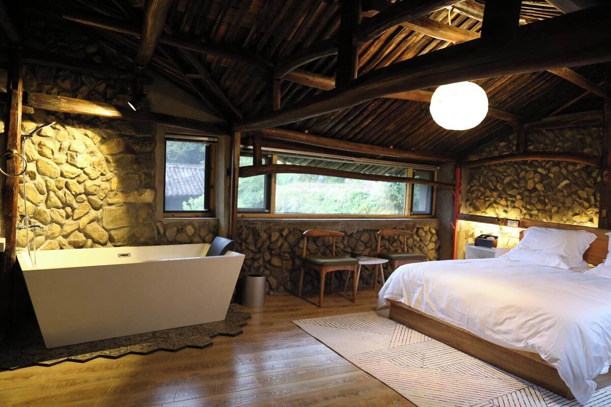 A room with a stone wall, a large bathtub, wooden flooring, and a bed with white bedding.
