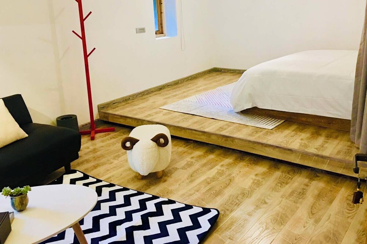 A bedroom with a bed, a small window, a red coat rack, a black and white rug, and a white fluffy animal decoration.