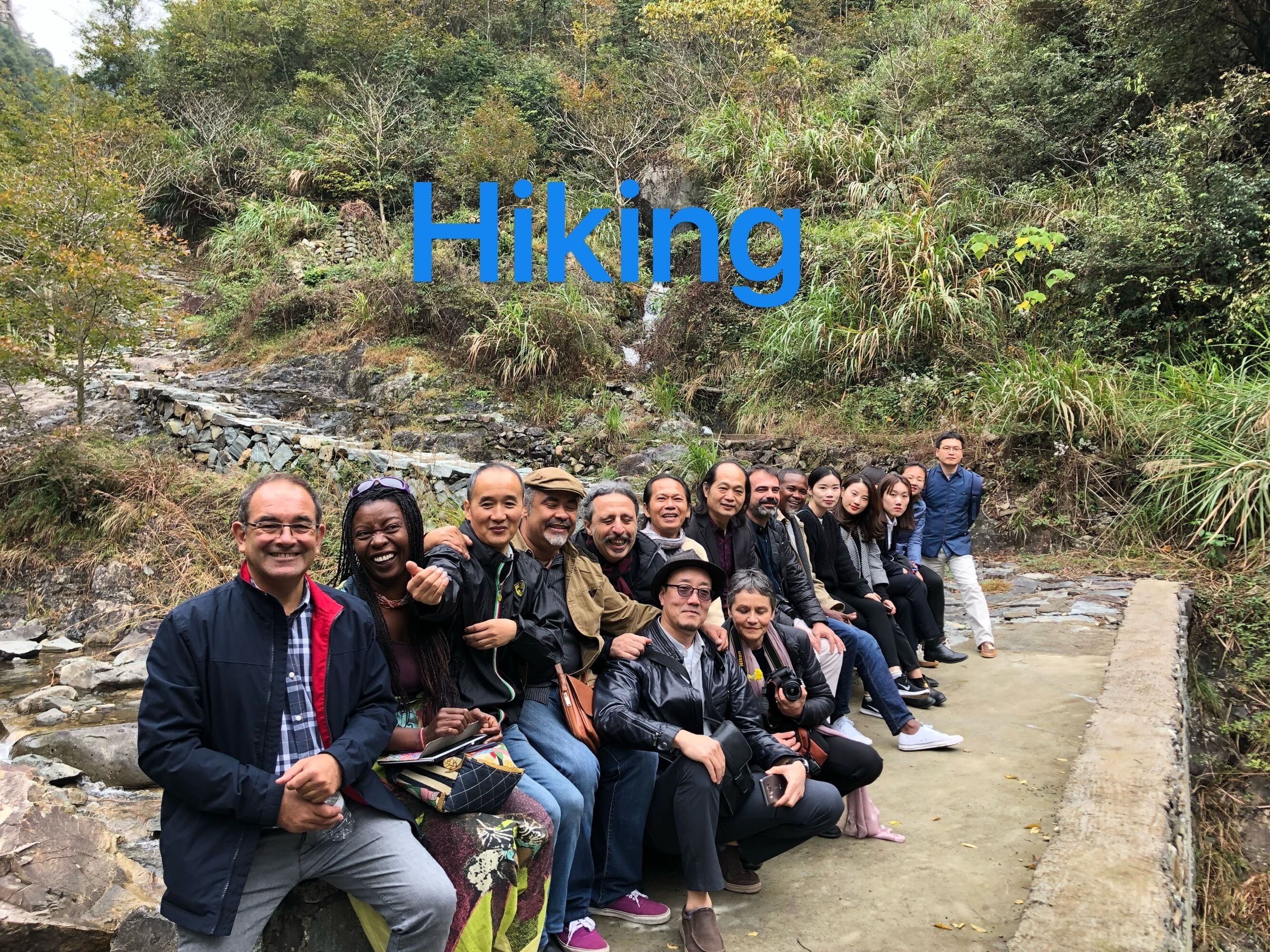 A group of people sitting on a concrete path surrounded by greenery, with the word "Hiking" overlaid.