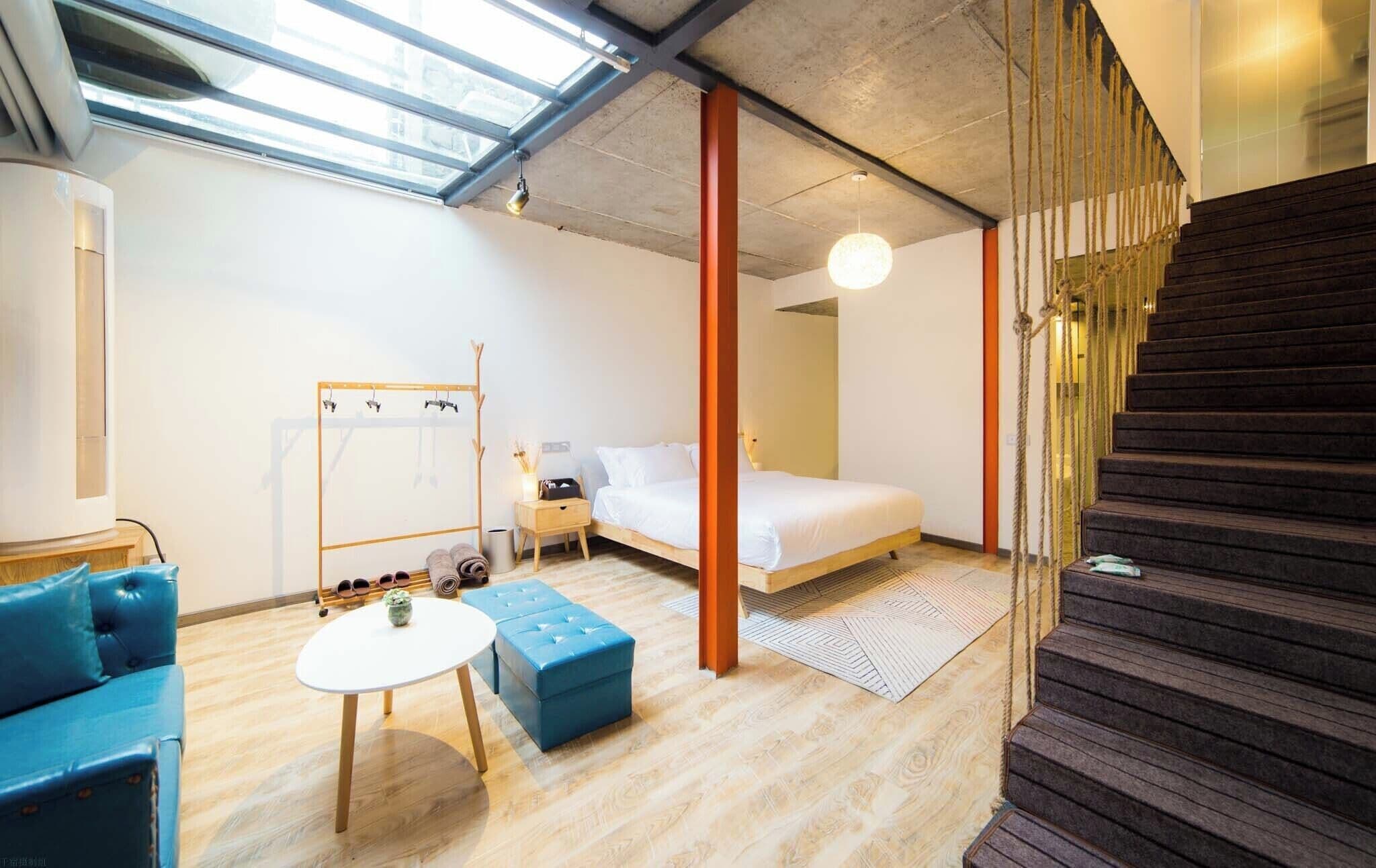A modern bedroom with a skylight, a bed, a small table, a blue ottoman, and a staircase.