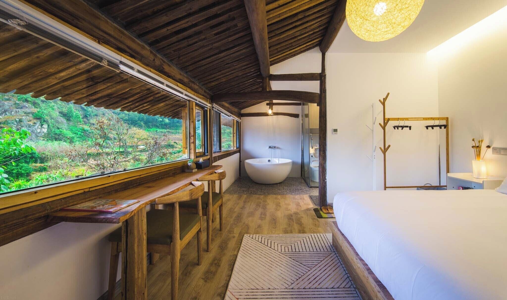 A room with a large window, a bathtub, a bed, and a wooden bench.