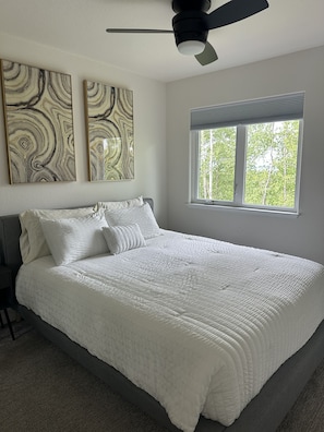 A bedroom with a bed, two wall-mounted artworks, a ceiling fan, and a window with a view of trees.