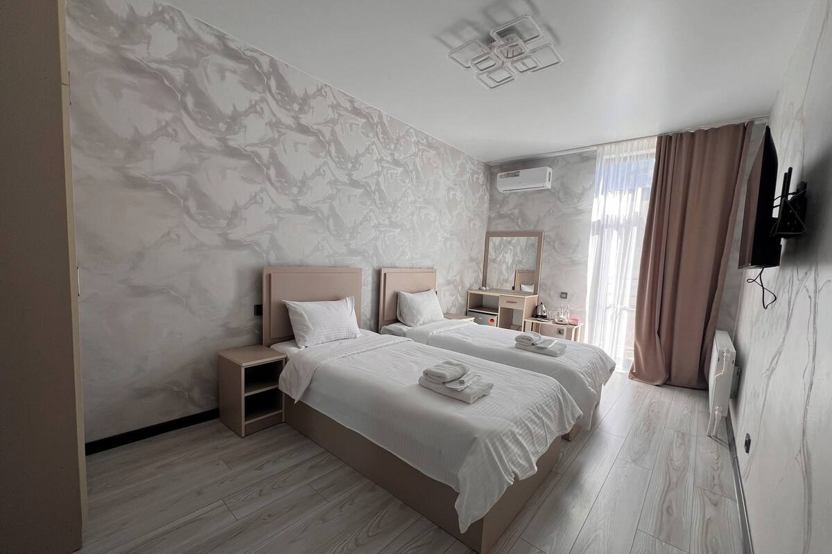 A modern hotel room with two beds, marble-patterned walls, a wooden floor, a flat-screen TV, and an air conditioning unit.