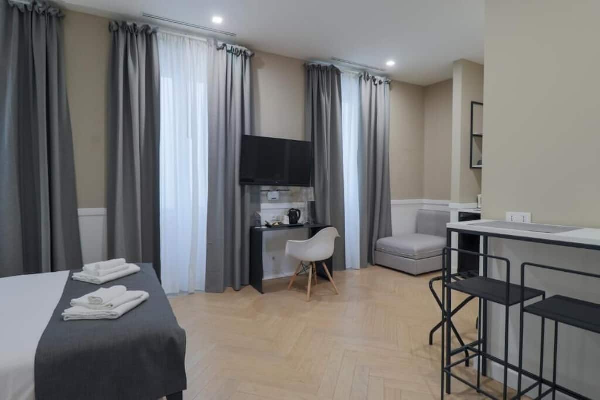 A hotel room with a bed, a desk with a chair, a TV, a small sofa, and a kitchenette area.
