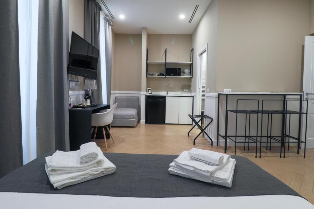 Habitación de hotel compacta con cama, escritorio, silla, kitchenette pequeña y televisor.
