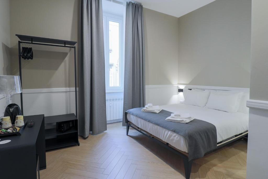 A modern hotel room with a bed, a desk with a lamp, a coat rack, and a window with curtains.