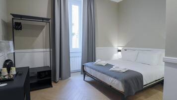 A modern hotel room with a bed, a desk with a lamp, a coat rack, and a window with curtains.
