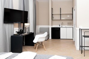 A modern hotel room with a flat-screen TV, a desk with a coffee maker, a white chair, a sofa, and a kitchenette with a sink and shelves. - Elisabeth Boutique Rooms (Trieste)