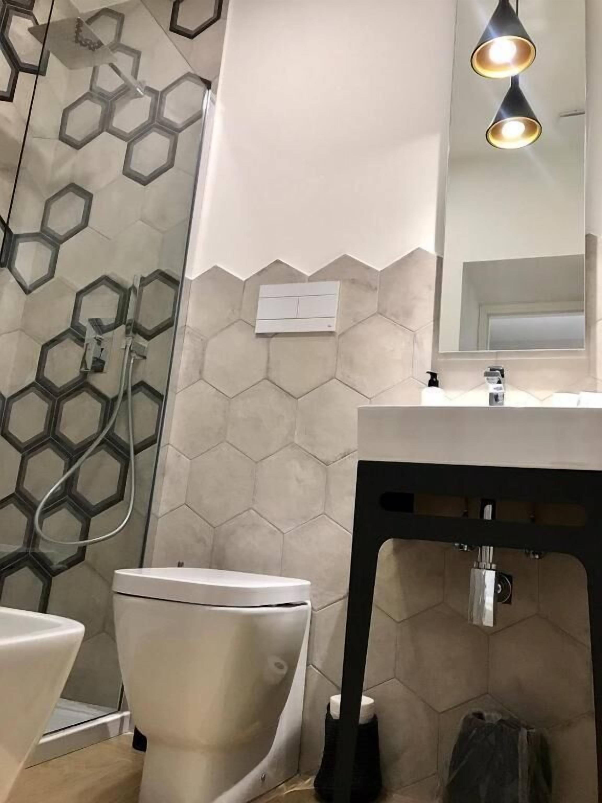 A modern bathroom with a hexagonal tile backsplash, a white toilet, a black sink with a mirror above, and a shower area with a glass door.