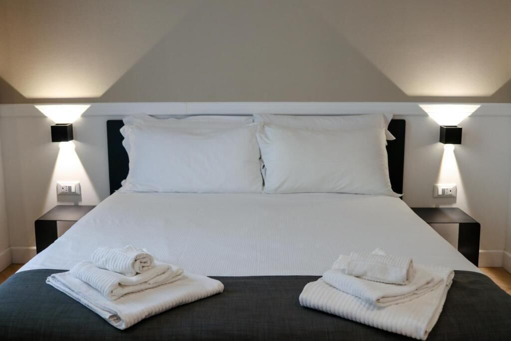 Una cama bien hecha con sábanas blancas, dos mesitas de noche con lámparas y toallas dobladas sobre la cama.