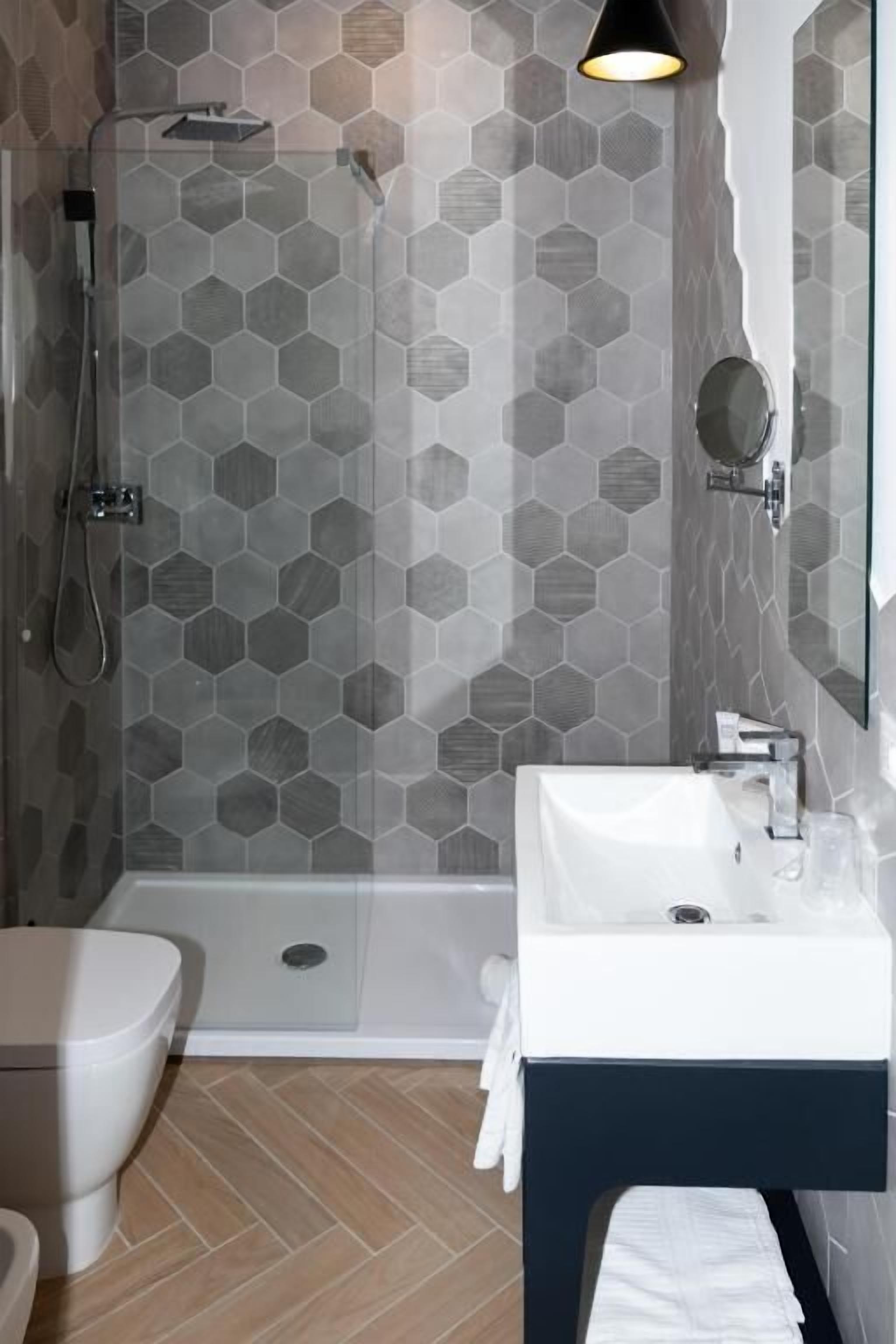 Un baño moderno con pared de ducha revestida de azulejos hexagonales, un lavamanos blanco con mueble oscuro y un inodoro.