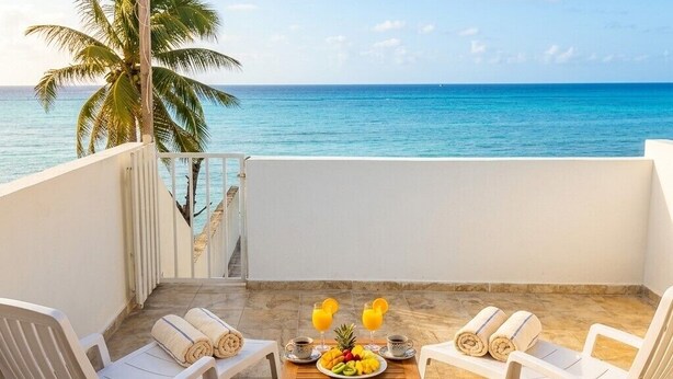 A balcony with two white chairs, a wooden table with a fruit platter, two glasses of orange juice, and a view of the ocean and palm tree. - MK Hotel Beach (Playa del Carmen)