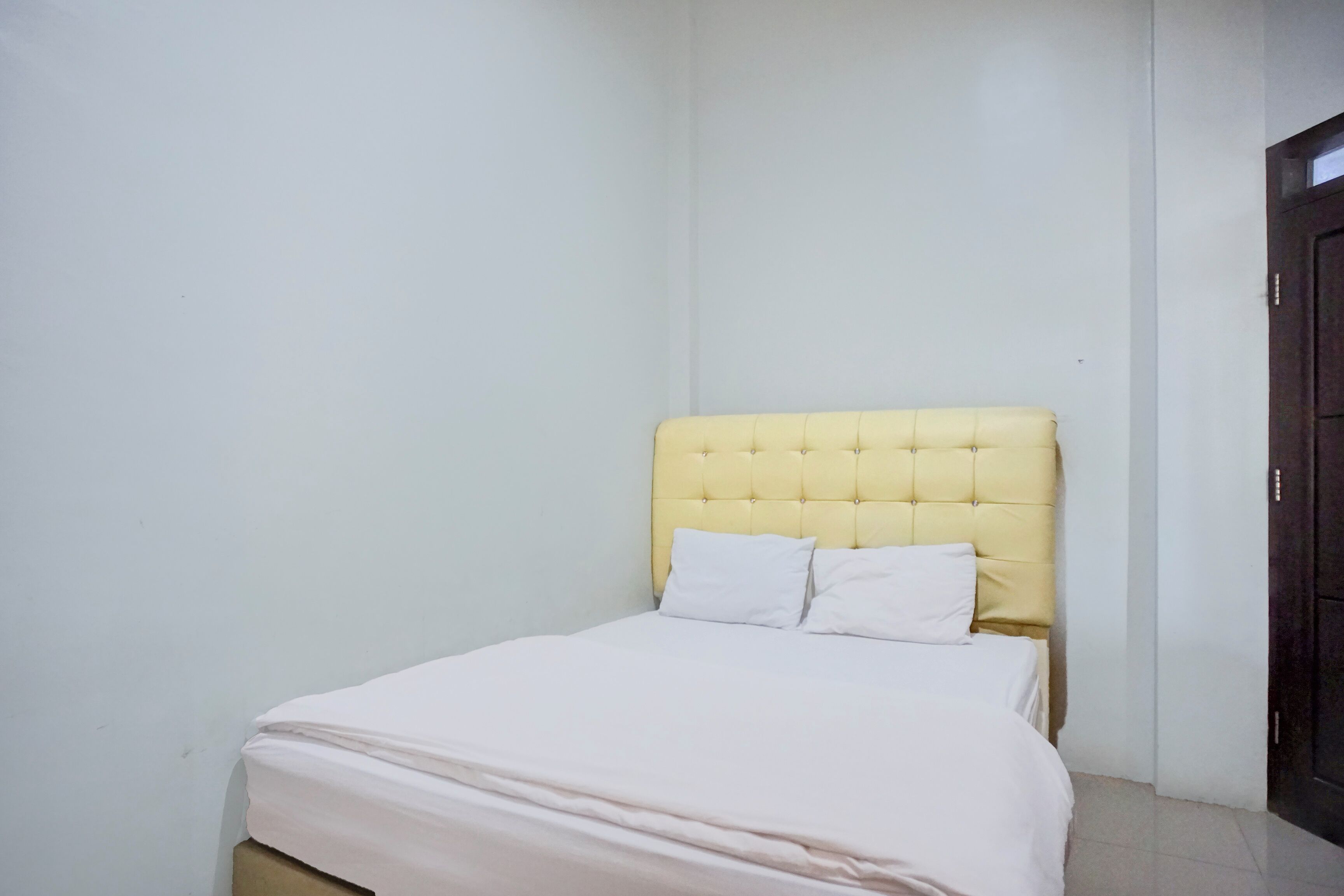 A small, neatly made bed with a yellow headboard and white bedding in a minimalistic room with plain walls and a dark door.