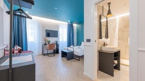A modern hotel room with a blue ceiling, a wooden floor, a white sofa, a desk with a computer, a small table, and a bathroom with a sink and mirror.