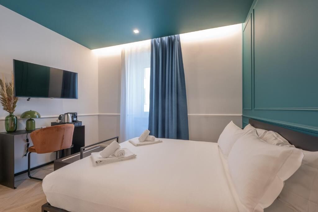 Een moderne hotelkamer met een groot bed, een flatscreen televisie aan de muur, een bureau met een stoel, en een raam met blauwe gordijnen.