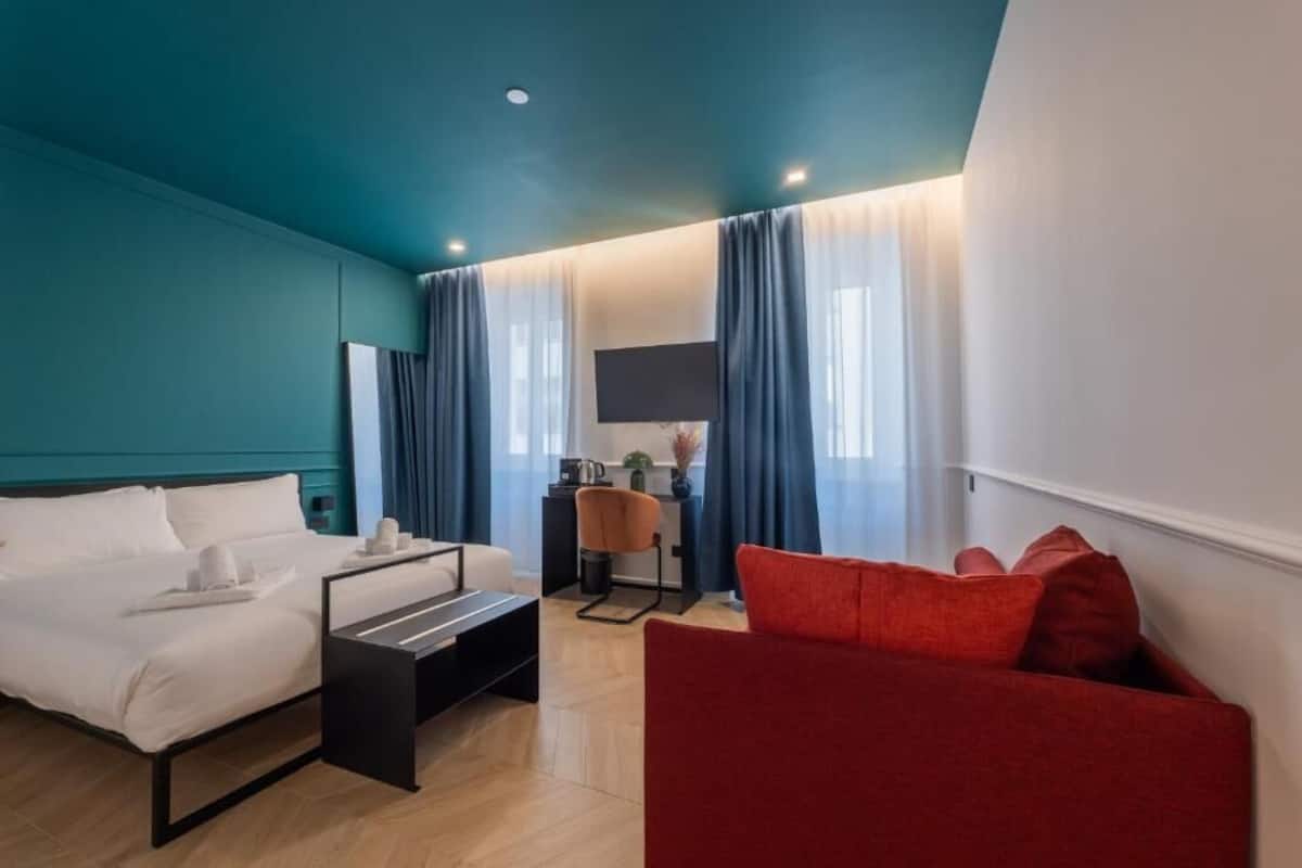 A modern hotel room with a teal accent wall, a bed with white linens, a red sofa, a small desk with a chair, and a flat-screen TV mounted on the wall.