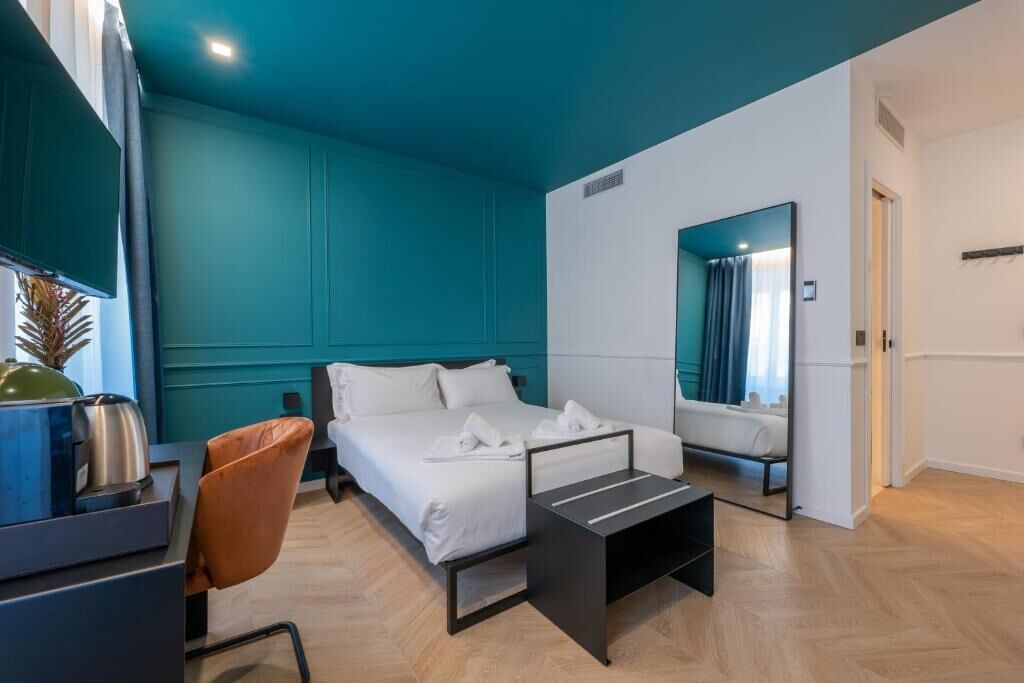 Una habitación de hotel moderna con paredes verde azulado, una cama con ropa blanca, un escritorio con una silla y una mesita.