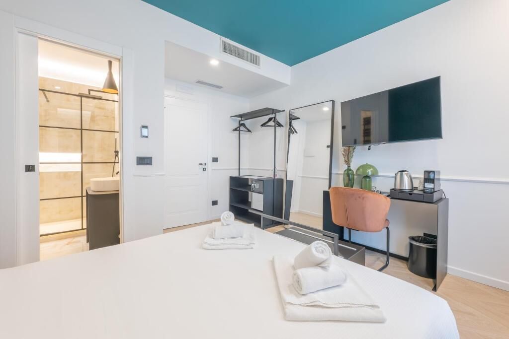 Quarto de hotel moderno com uma cama grande, televisão de tela plana, espelho, pequena mesa com cafeteira e banheiro visível através de uma porta aberta.