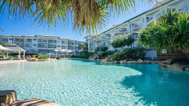 A clear swimming pool with lounge chairs and umbrellas, surrounded by a resort building with multiple balconies. - The Pool Resort Kingscliff - One Bedroom (7302) - Holiday Management Co. (Tweed)