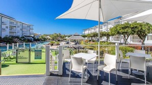 An outdoor seating area with white tables and chairs, a large umbrella, and a view of a swimming pool and buildings in the background. - The Pool Resort Kingscliff - One Bedroom (7302) - Holiday Management Co. (Tweed)