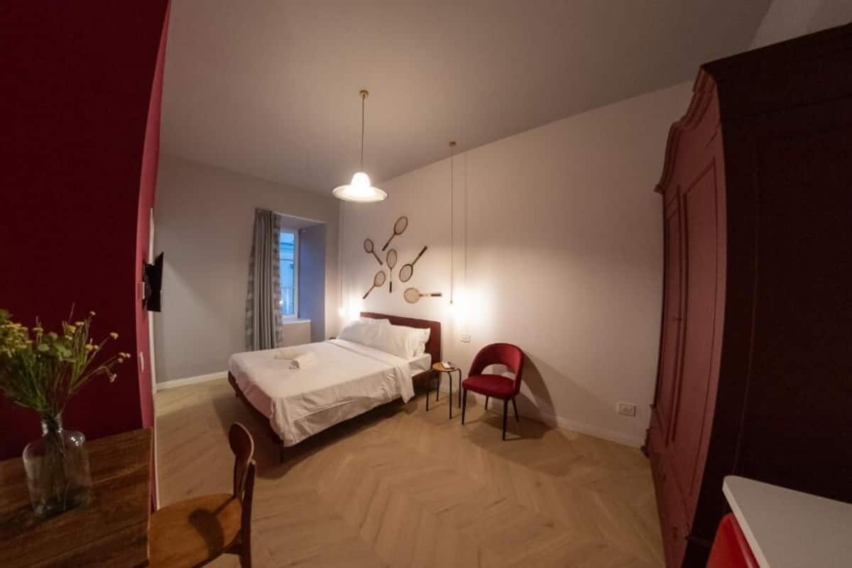 A bedroom with a bed, a chair, a small table, and a wall-mounted light fixture.