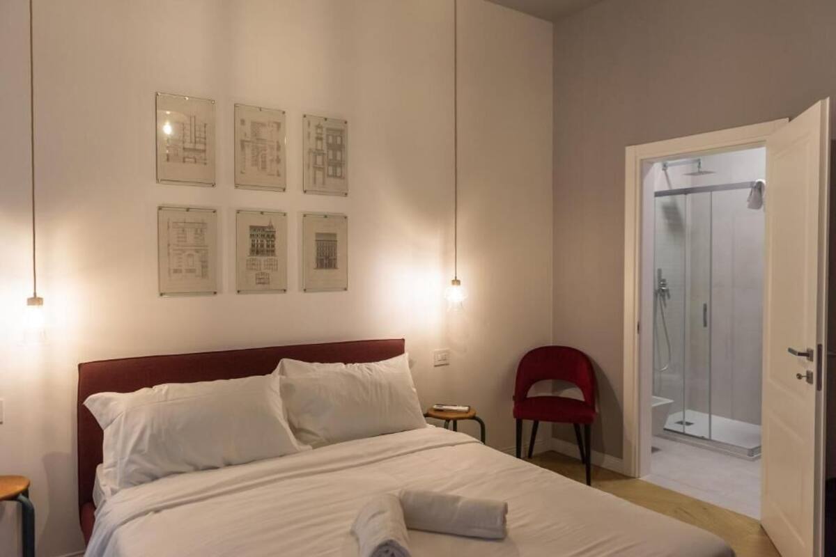 A neatly made bed with white linens, a red headboard, and a small bedside table with a lamp. There are framed pictures on the wall and a red chair near the door.