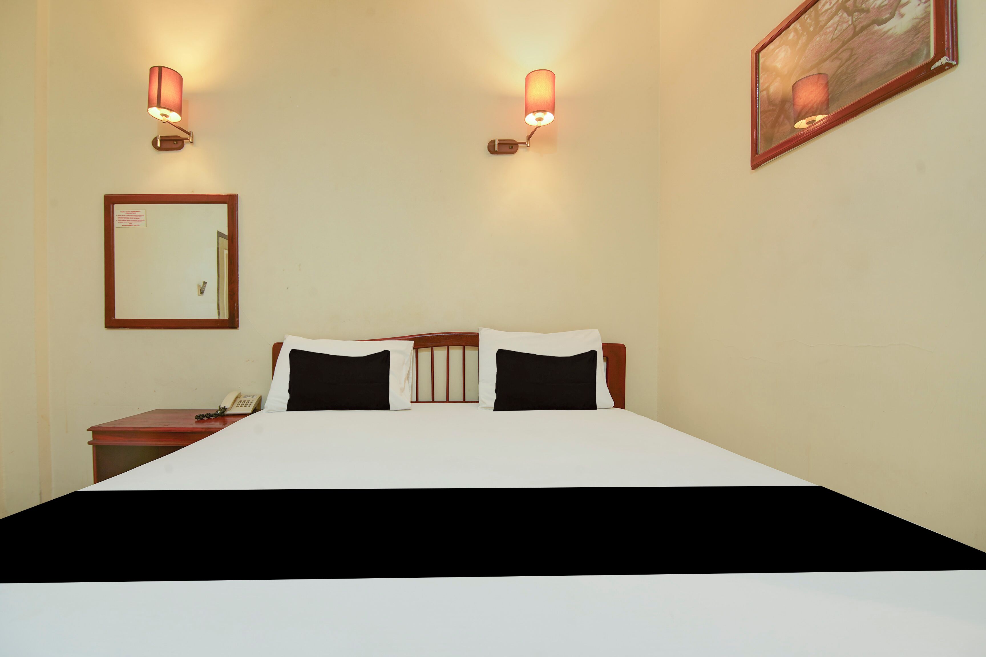 A hotel room with a large bed, two bedside lamps, a nightstand with a phone, a mirror, and a painting on the wall.