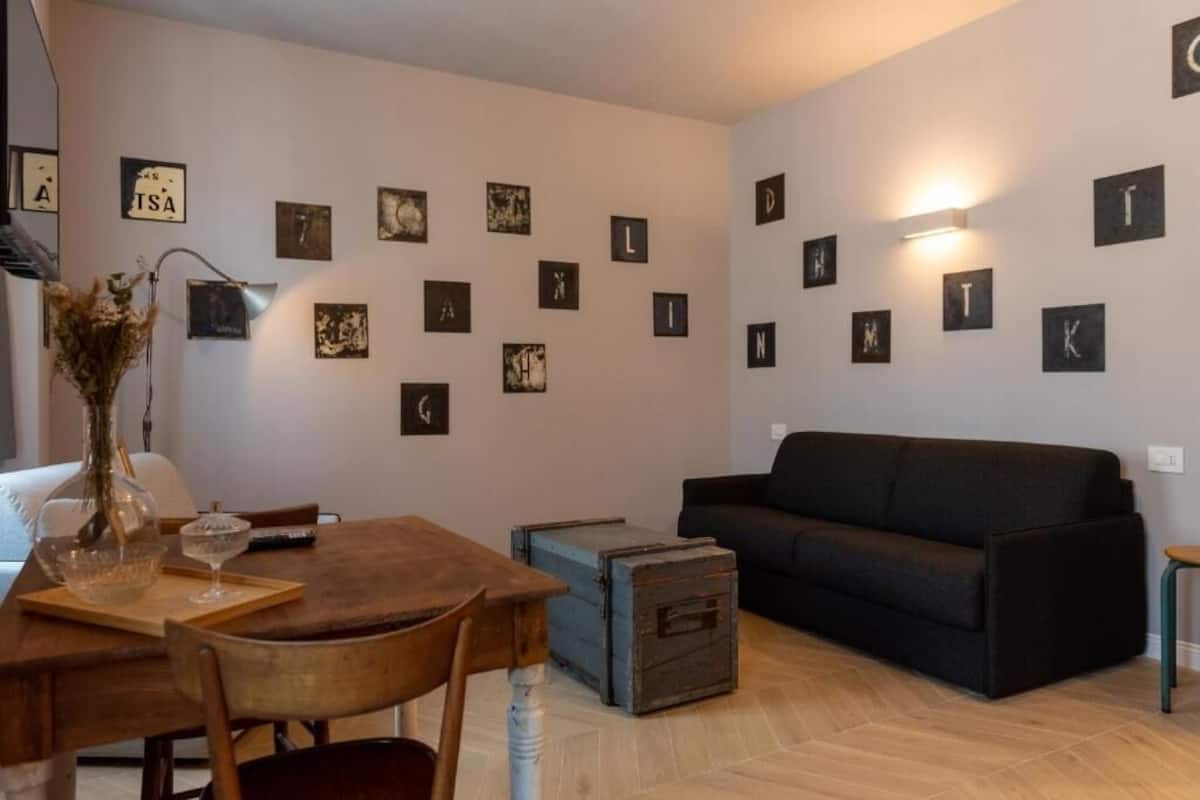 A living room with a dark sofa, a wooden dining table with chairs, and a vase with dried flowers. There are framed pictures on the walls and a small blue chest on the floor.