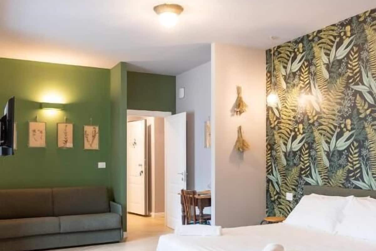A hotel room with a bed, a sofa, a dining table, and a wall with a leafy pattern.