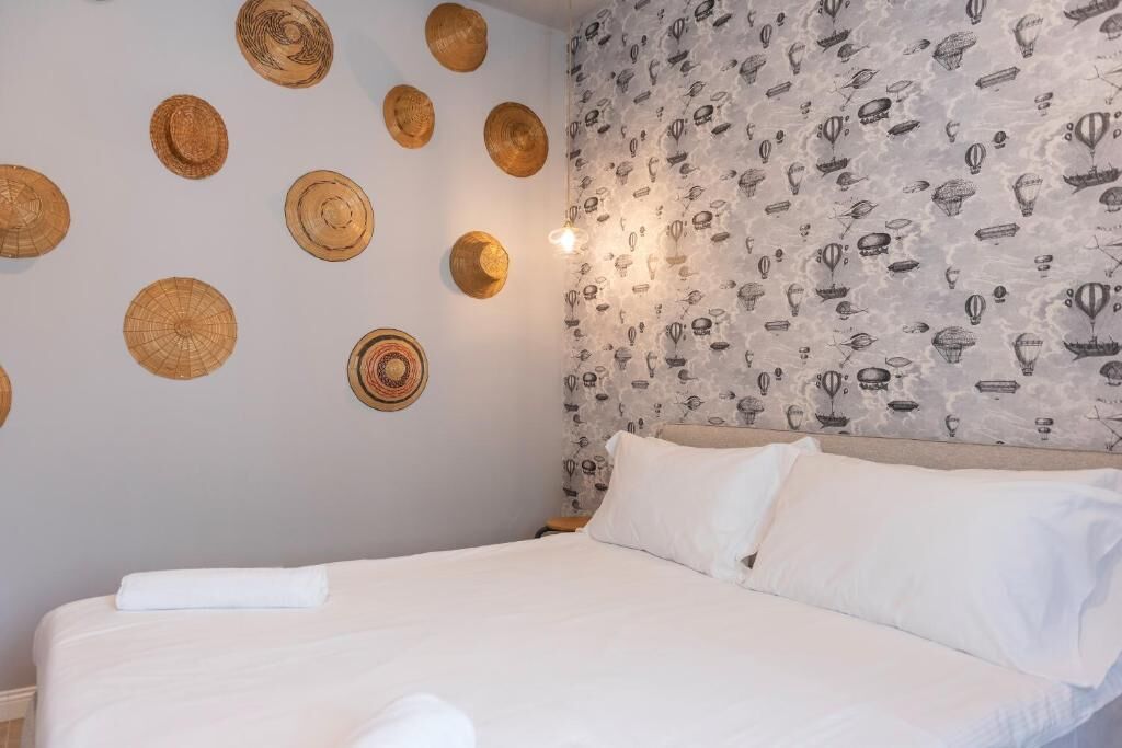 Ein ordentlich gemachtes Bett mit weißen Leinen und Kissen, eine Wand mit gewebten Hüten und Tapete mit verschiedenen Motiven.
