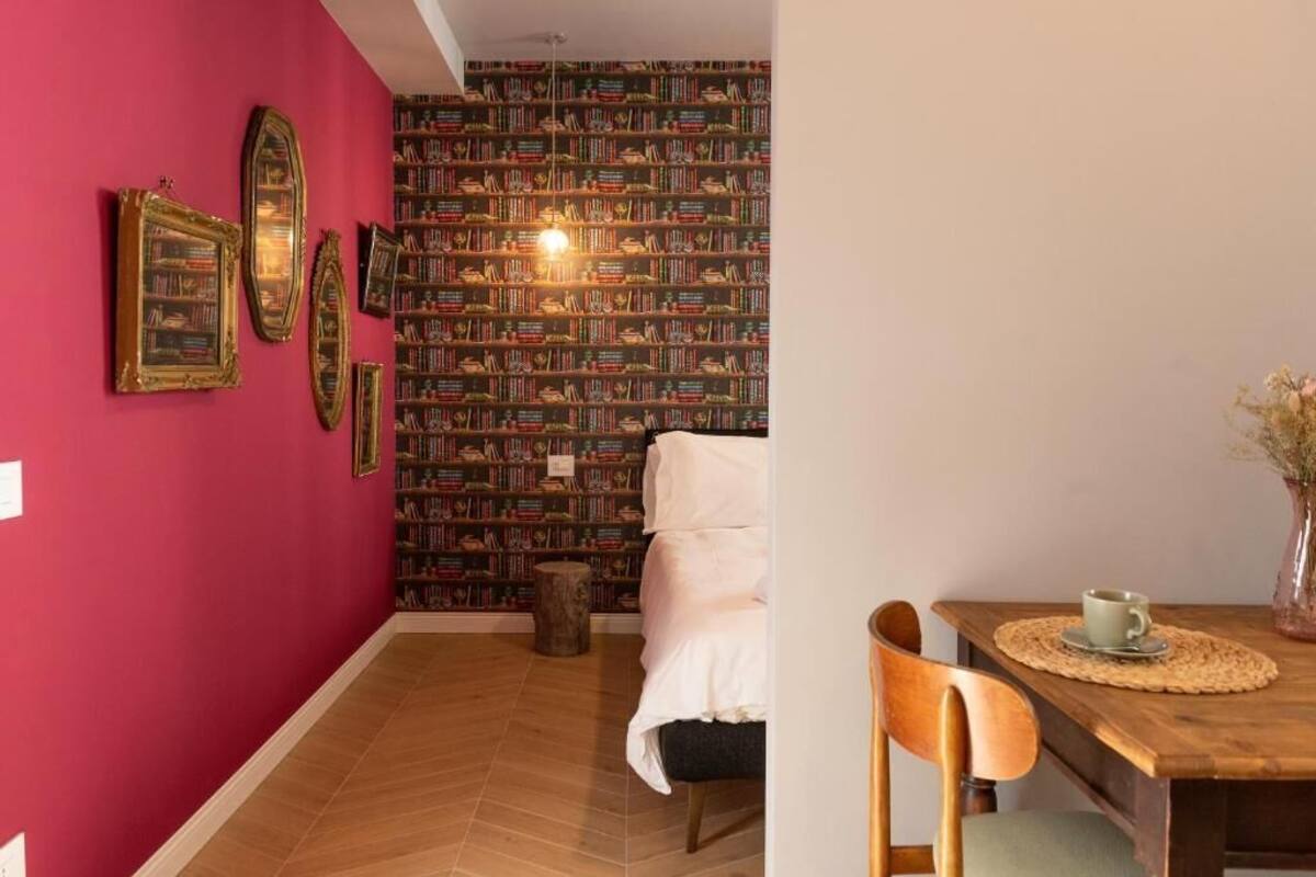 A room with a red wall, a bookshelf, a bed, and a wooden dining table with chairs.
