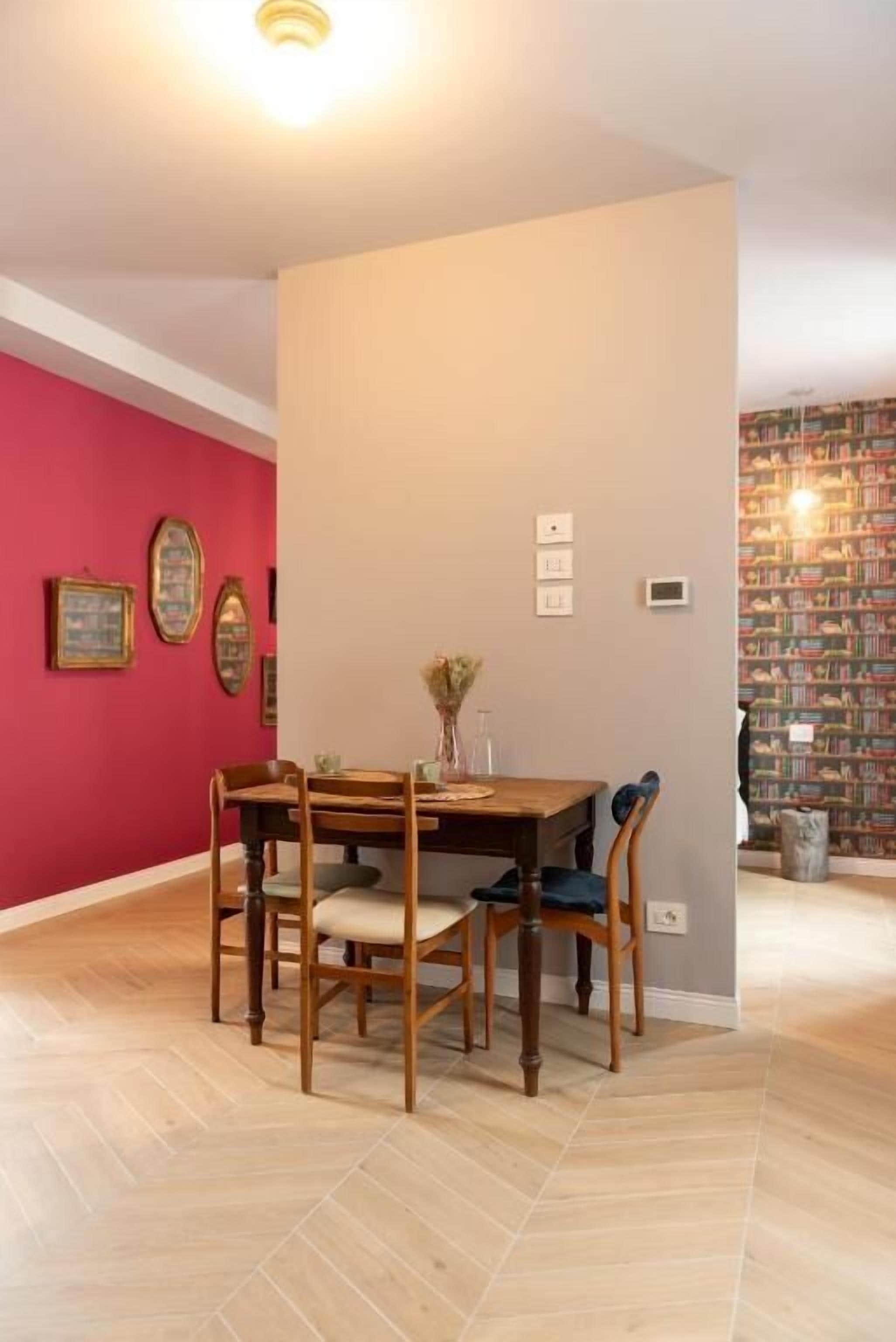 A dining area with a wooden table and chairs, a red accent wall, and a bookshelf in the background.