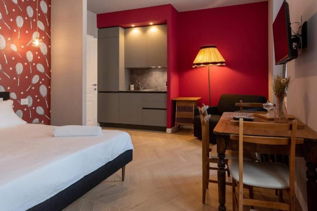 A hotel room with a bed, a dining table with chairs, a lamp, and a kitchenette in the background.