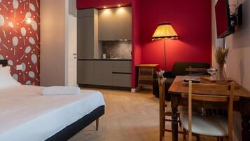 A hotel room with a bed, a dining table with chairs, a lamp, and a kitchenette in the background.