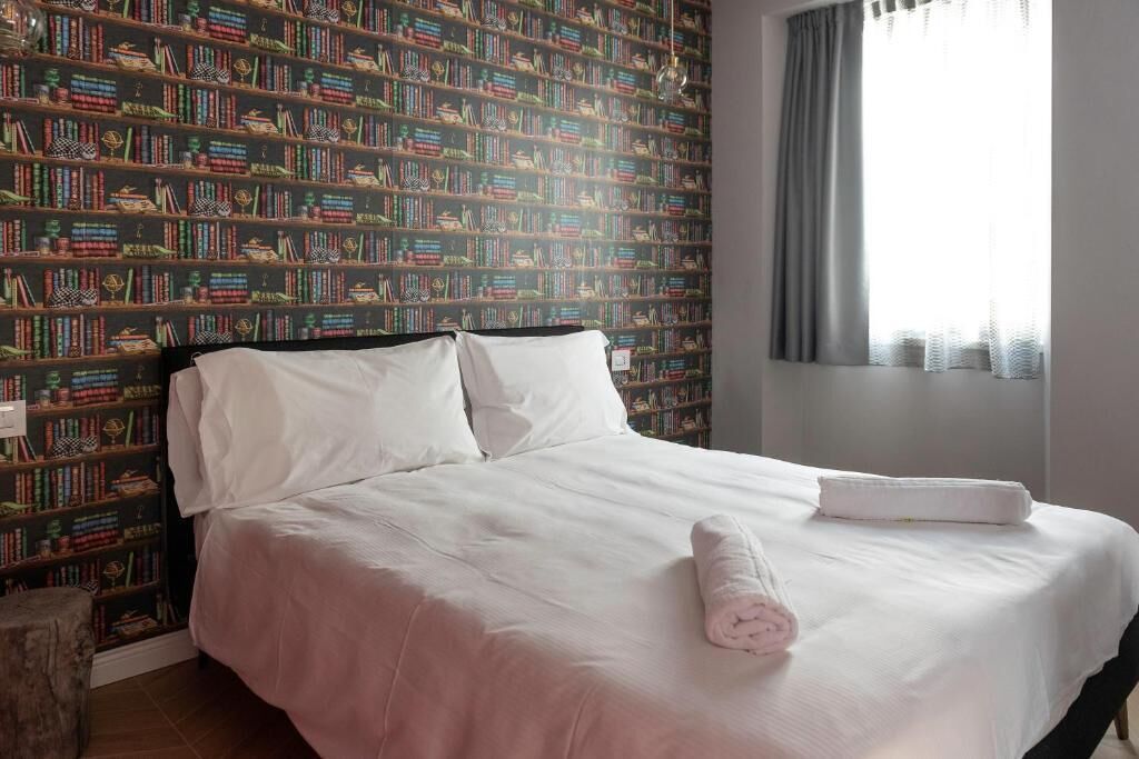 Una cama bien hecha con sábanas blancas y una toalla enrollada, una estantería llena de libros coloridos y una ventana con cortinas.