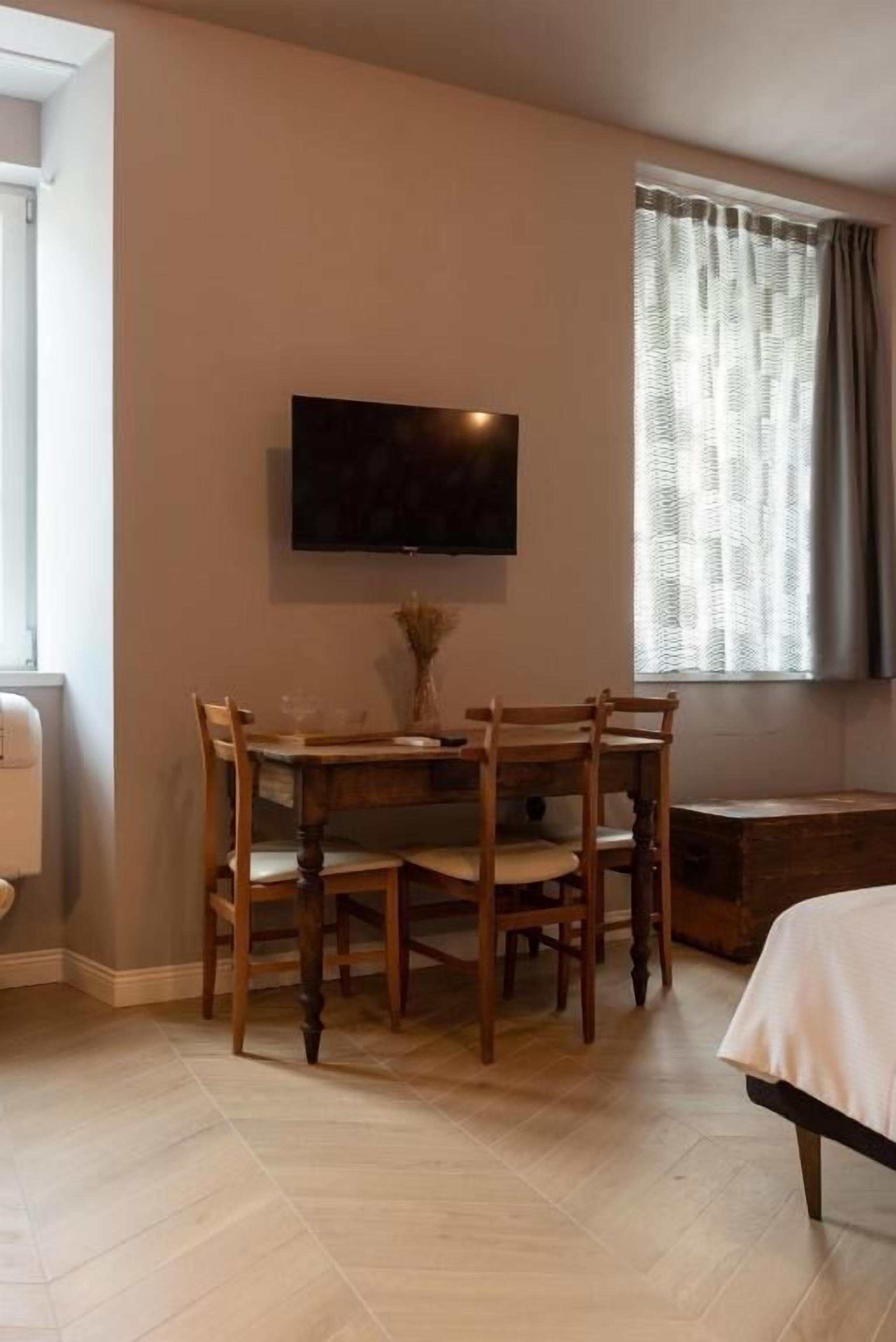 A room with a wooden dining table, four chairs, a flat-screen TV mounted on the wall, a window with curtains, and a bed with a white bedspread.
