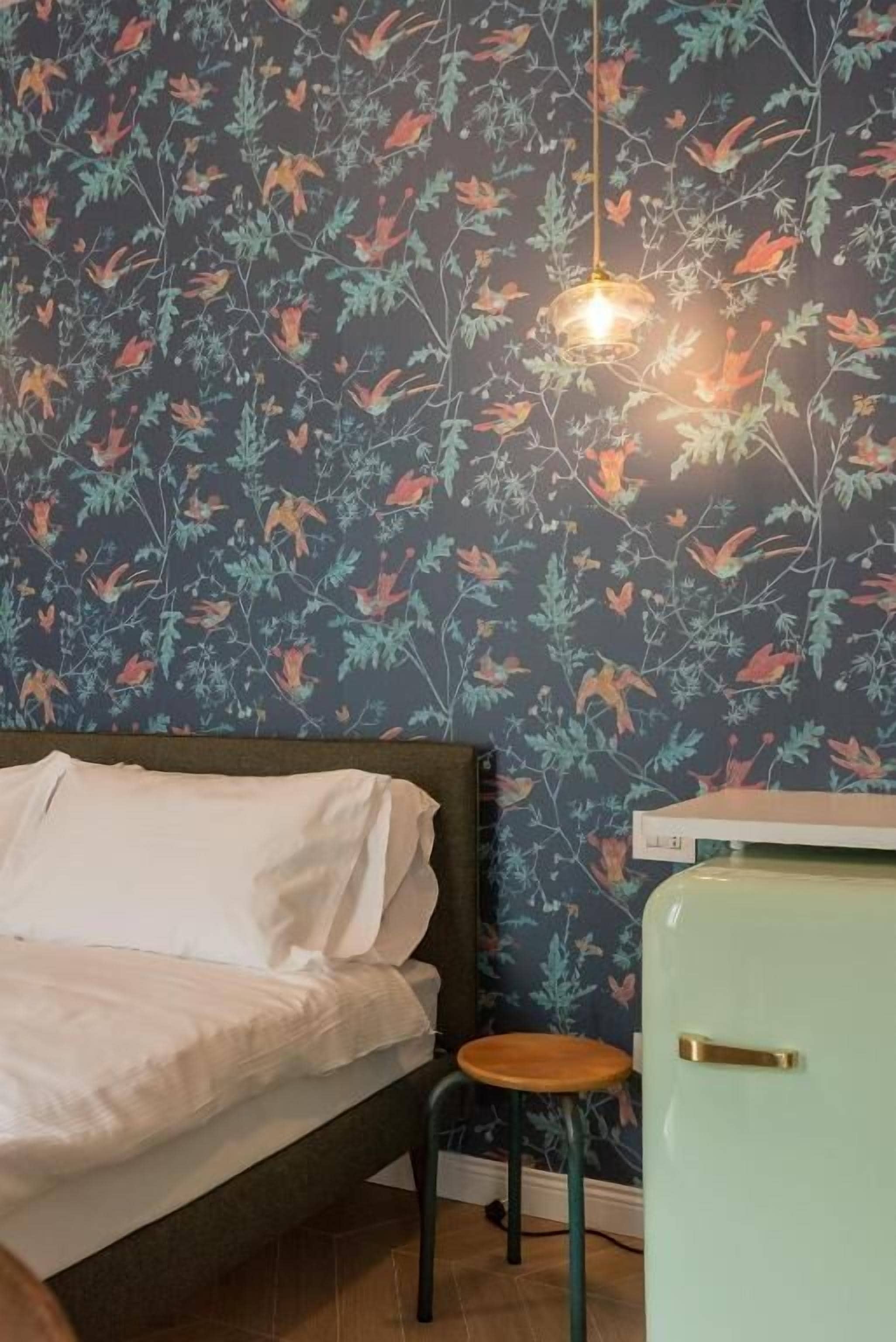Ein Zimmer mit einem Bett, einem kleinen Hocker, einem Kühlgerät im Vintage-Stil und floralem Wandpapier.