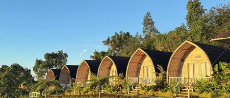 A row of wooden, arched-roof cottages with a grassy hill and trees in the background.
