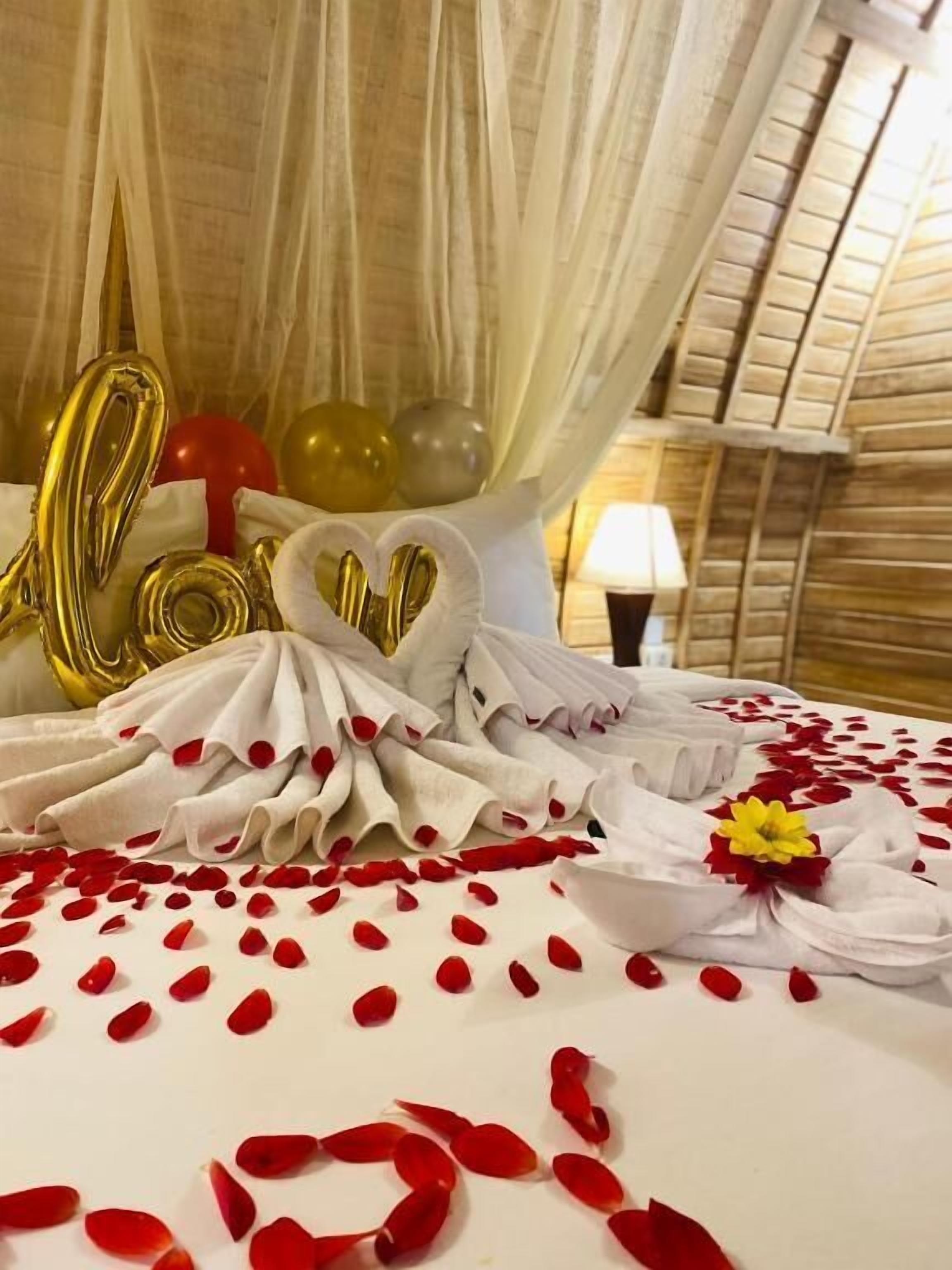 Una cama con sábanas blancas, pétalos de rosa roja y globos dorados que deletrean 'LOVE'.